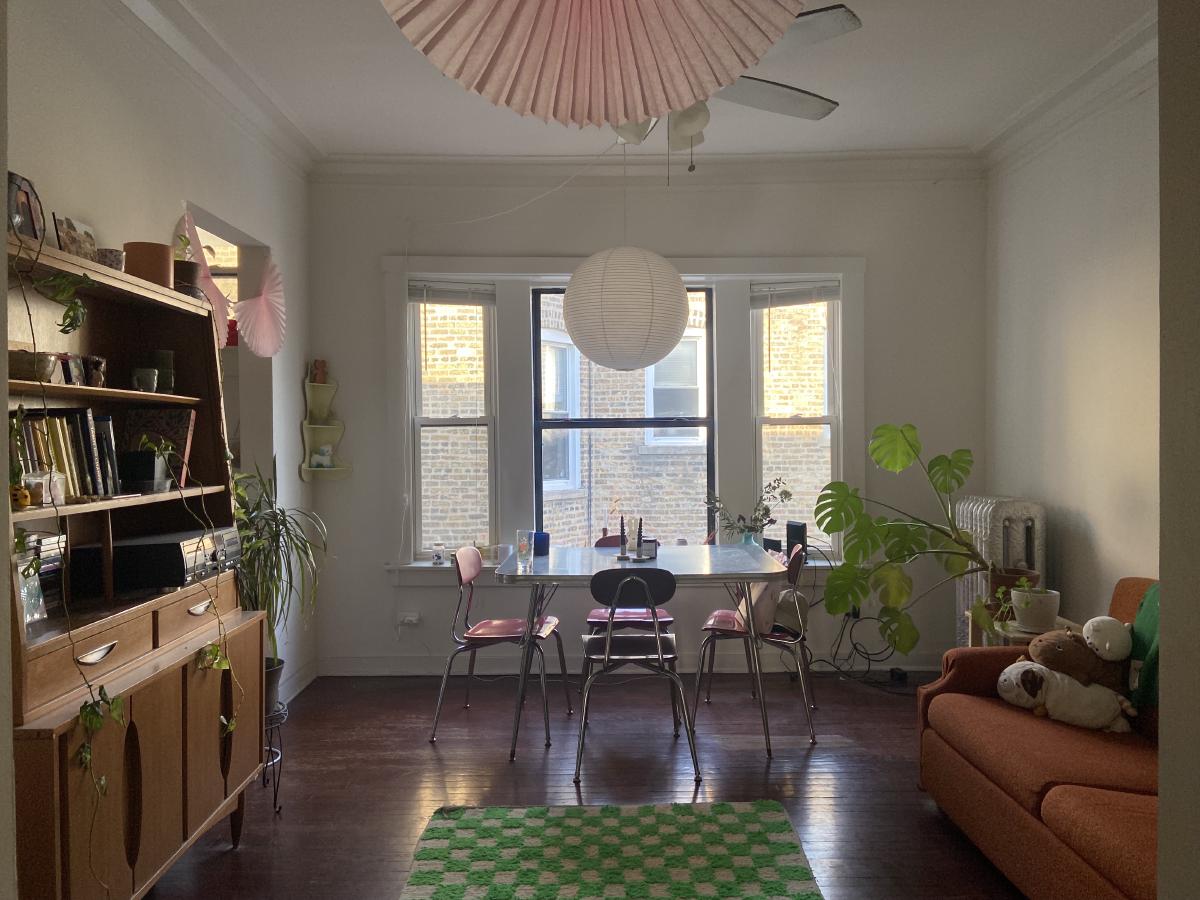 our kitchen with a green checkered rug table with red chairs wooden bookshelf orange couch with push and plants